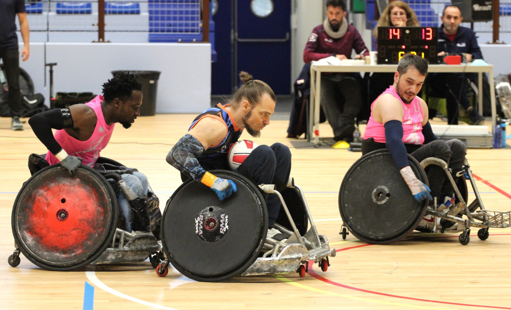 trois joueurs de rugby fauteuil en action sur le parquet, table de marque en arrière plan